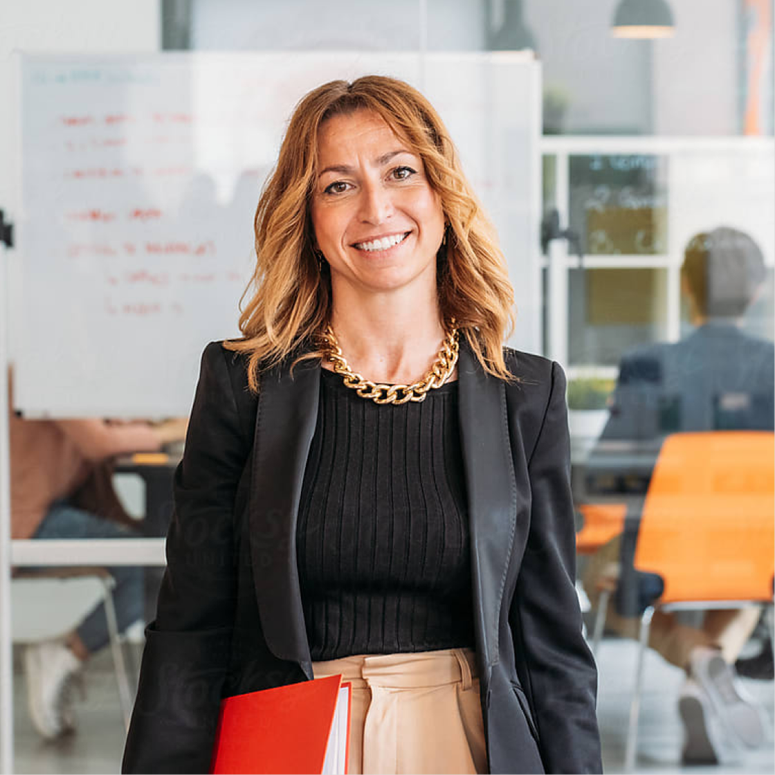 Image of a woman standing, wearing a blazer, holding a red notebook, and smiling in an office setting