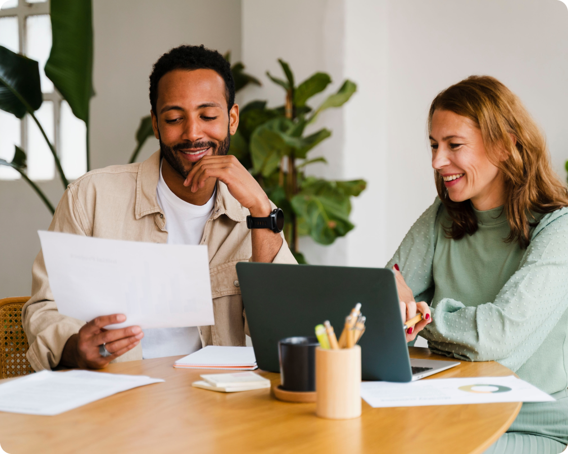 Image of a man wearing a tan shirt holding a piece of paper with a woman wearing a teal shirt looking at a computer