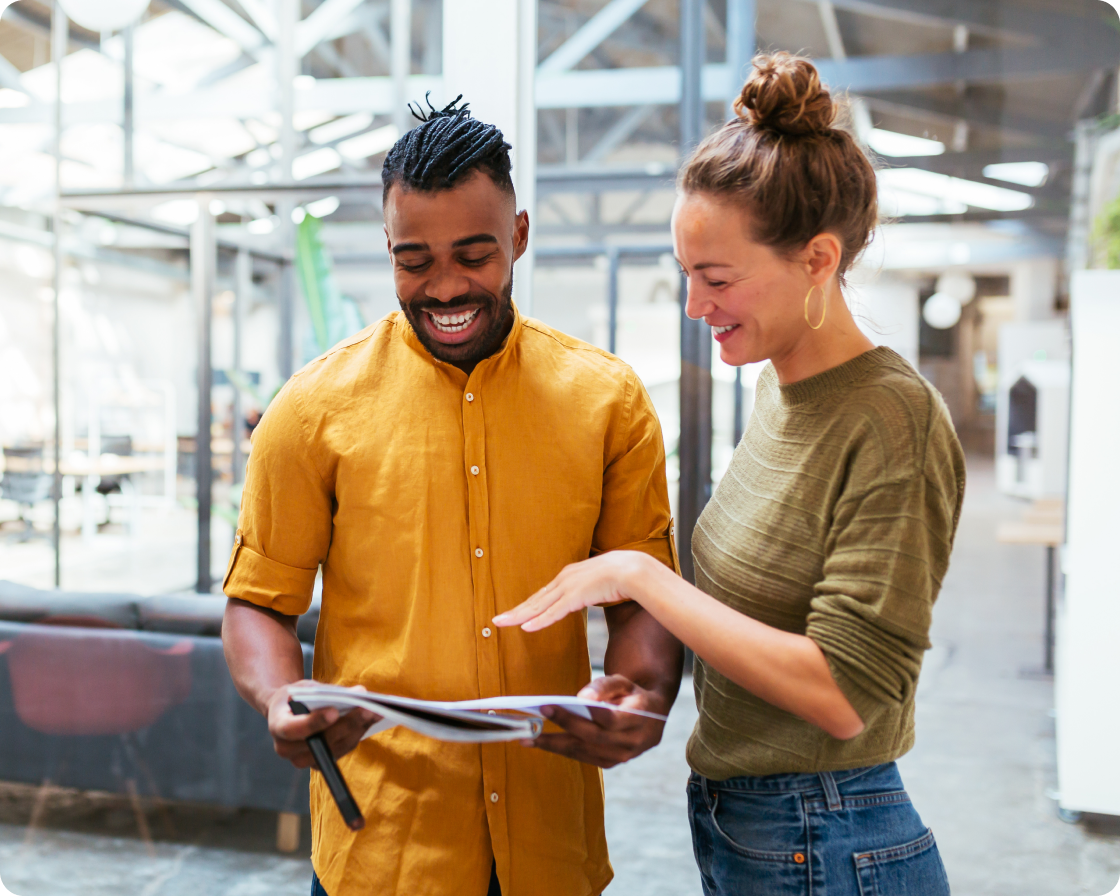 Image of a man wearing a yellow shirt holding papers, with a woman wearing a green shirt standing and looking at papers in an office setting