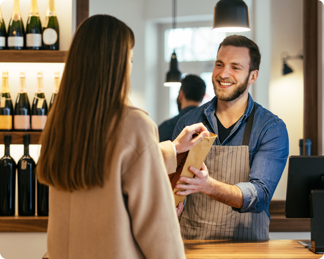 Image of a man in a tan apron and blue shirt working at a store handing a package to a female customer