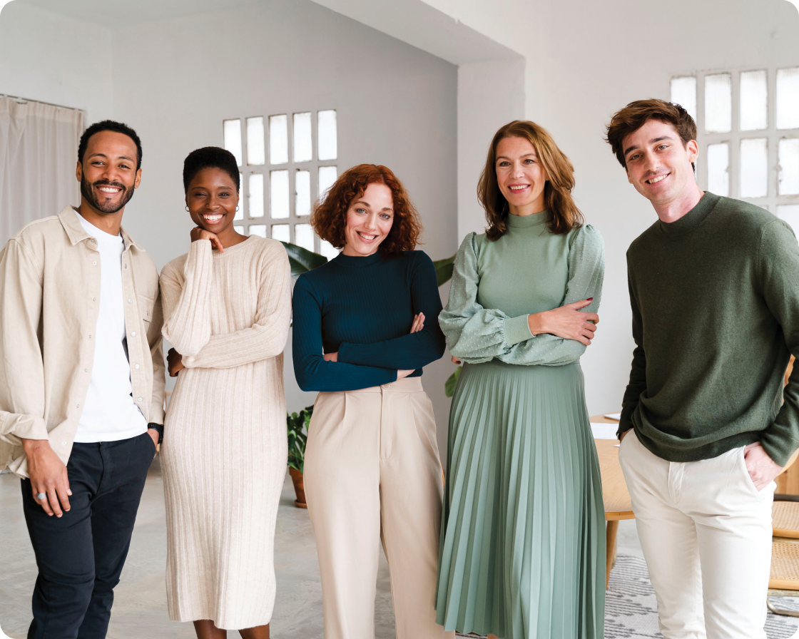 Image of five people standing in a line in various poses wearing cream colors, blues, and green shades of clothing smiling to camera in a white office setting