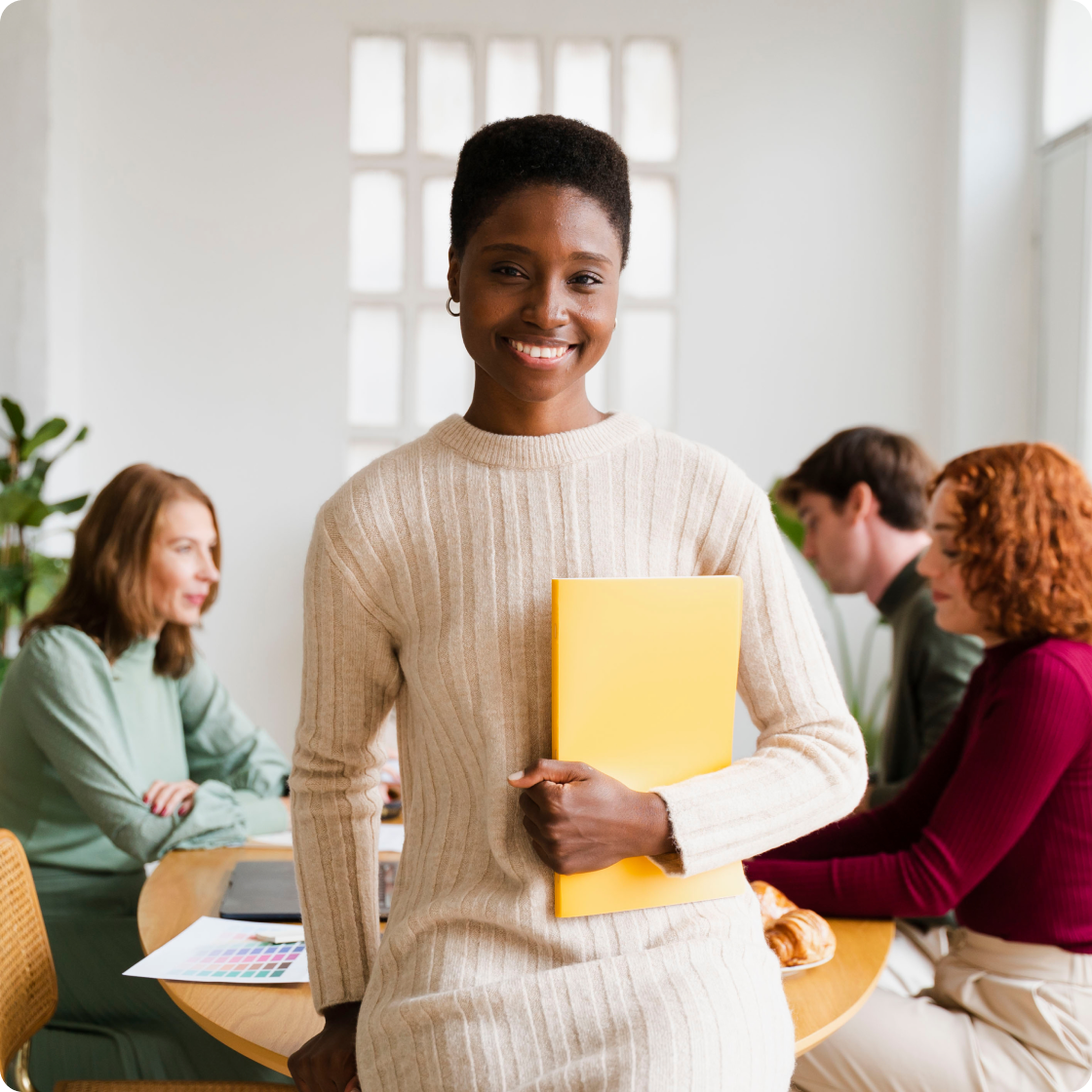 Image of a woman smiling to camera, leaning on a conference table, holding a yellow folder