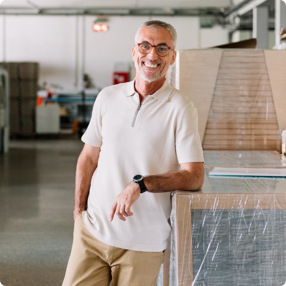 Image of a man in a white shirt and tan pants smiling and leaning against a pallet in a warehouse