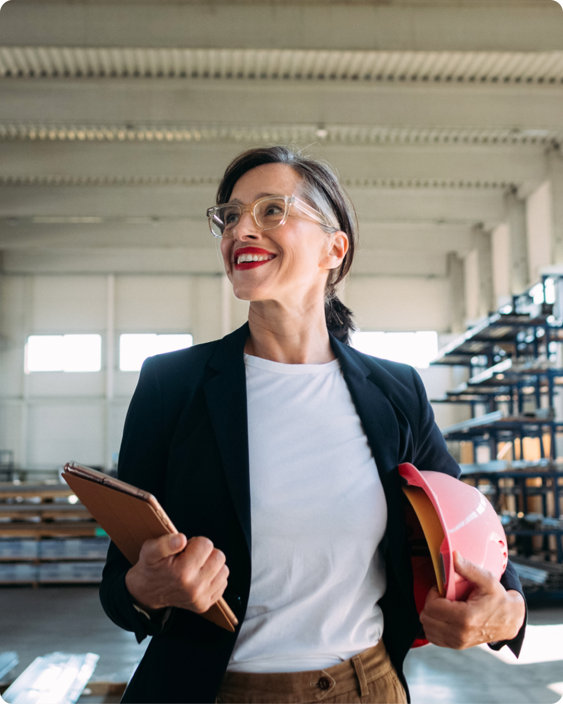 Woman wearing a blue blazer and white shirt holding a hat and a clipboard smiling in a warehouse setting