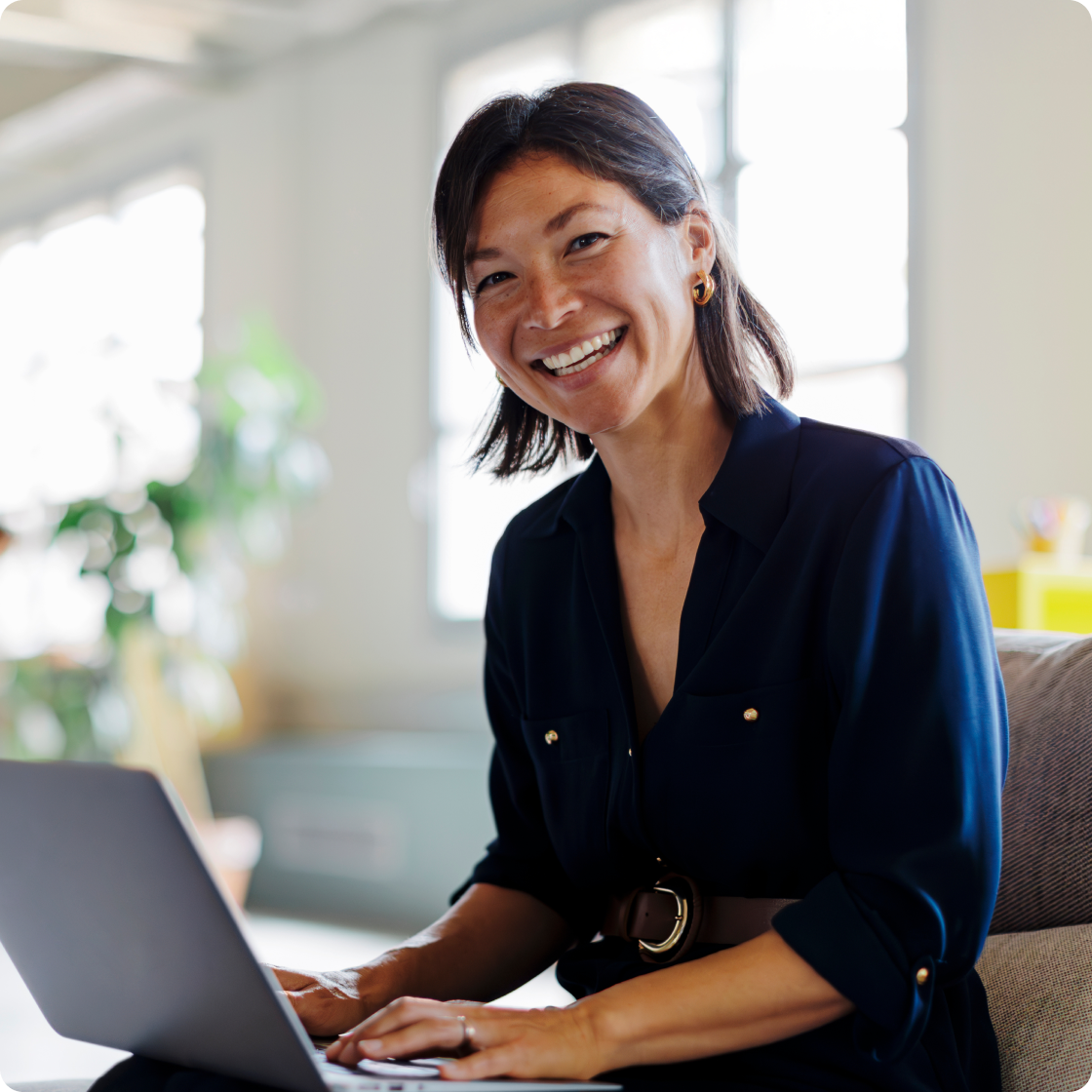 Woman wearing a blue blouse sitting a desk typing on her computer smiling to camera