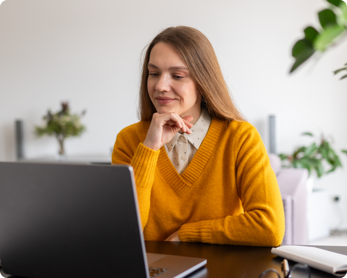 Woman at desk with her head leaning on her hand smiling down at a laptop.