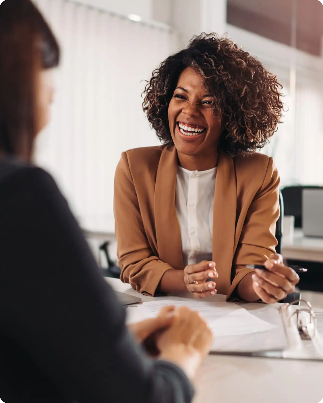 Woman smiling at a desk talking to another woman in an office setting.